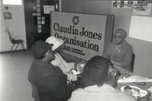black-and-white photograph showing a group of four women sitting at a tableBlack-and-white photograph showing a group of four women sitting at a table. On the wall behind them, a large banner reads "Claudia Jones Organisation (An Afro-Caribbean Women's Organisation)." One of the women, leading the discussion, sits facing the other three who have their backs to the camera. The setting appears to be informal, with a table covered in a patterned cloth, papers, and a plate of food visible.